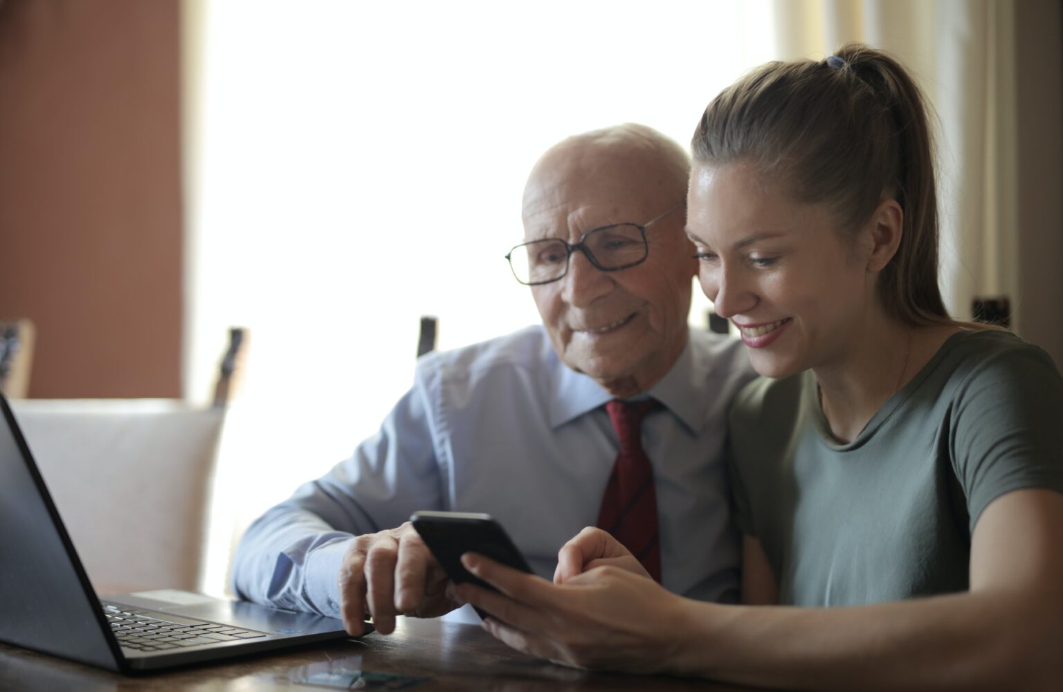 Persona mayor usando un smartphone con ayuda de una mujer más joven, ambos sentados frente a un ordenador, simbolizando el apoyo familiar y el uso de tecnología en el cuidado domiciliario.